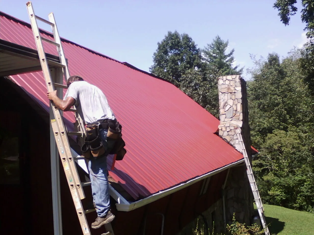 Roofer on a ladder beside a red metal roof and stone chimney — Soffit & Fascia Repair by Roofing Brothers in Glen Rock